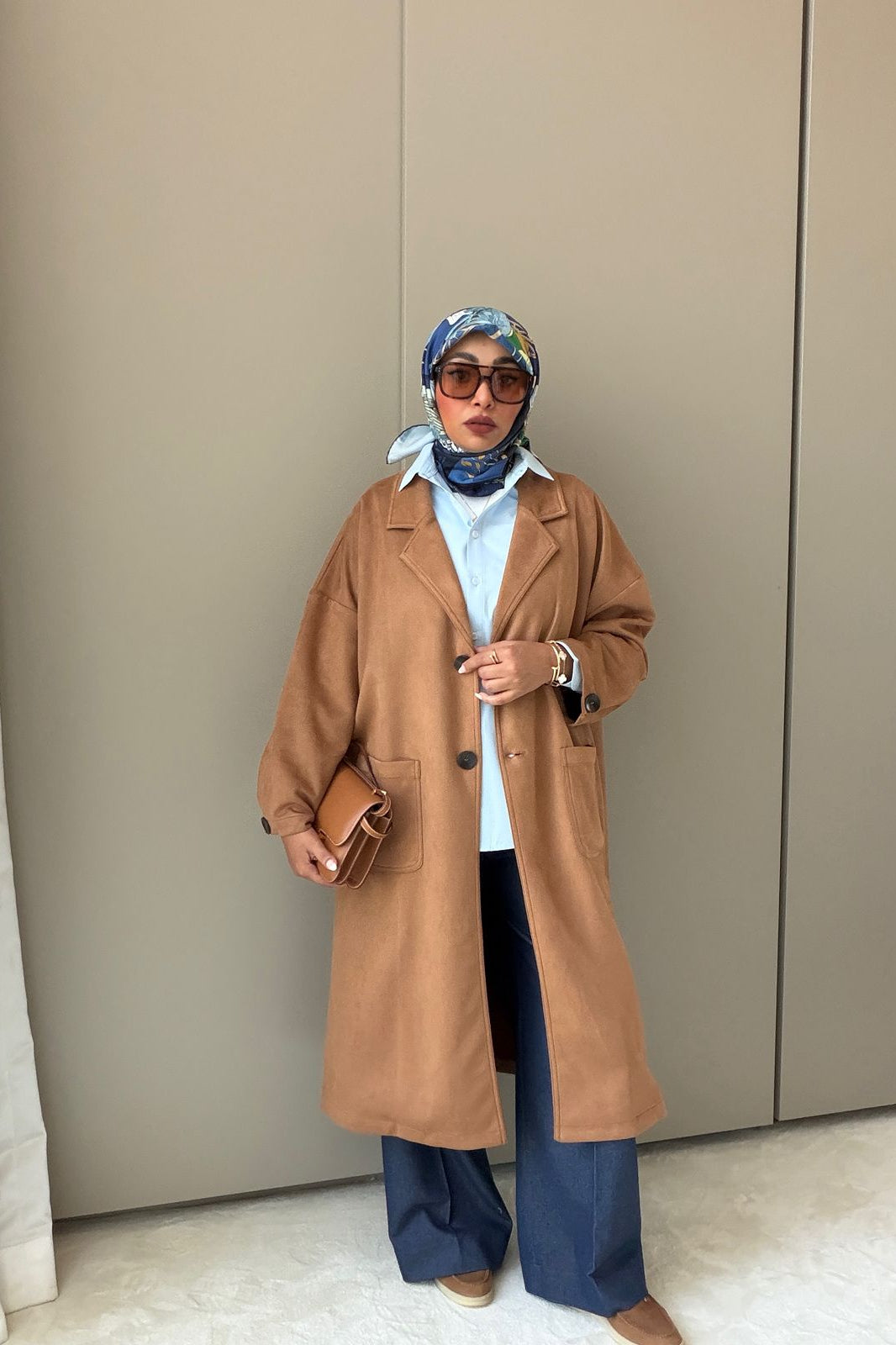 Woman wearing a long brown coat, blue pants, and a headscarf against a beige wall.