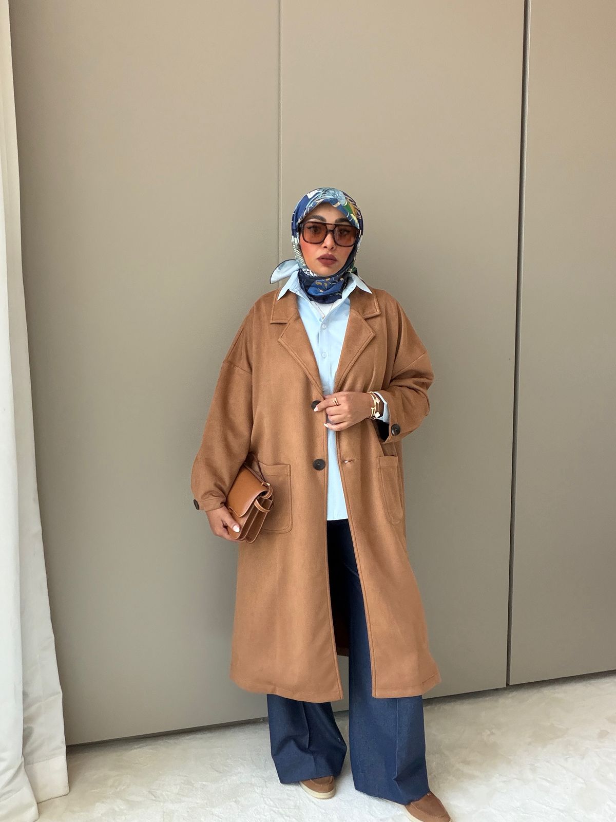 Woman wearing a long brown coat, blue pants, and a headscarf against a beige wall.