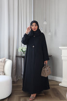 A person wearing a black abaya with a scarf, standing in a room with light decor.